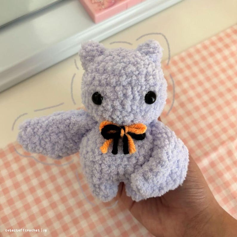 May include: A crocheted purple bat with orange and black ribbon. The bat has black eyes and a fluffy texture.