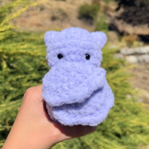 May include: A small, crocheted purple hippopotamus toy with black eyes. The toy is being held in a person's hand.