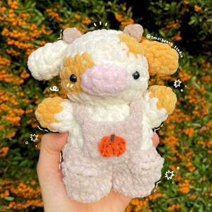 May include: A crocheted plush toy cow with a white, pink, and orange color scheme. The cow is wearing overalls with a pumpkin patch design. The toy is held in a hand.
