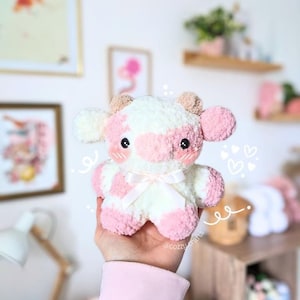 May include: A pink and white crocheted cow plush toy with a white bow. The toy is being held in a hand wearing a pink sweater.