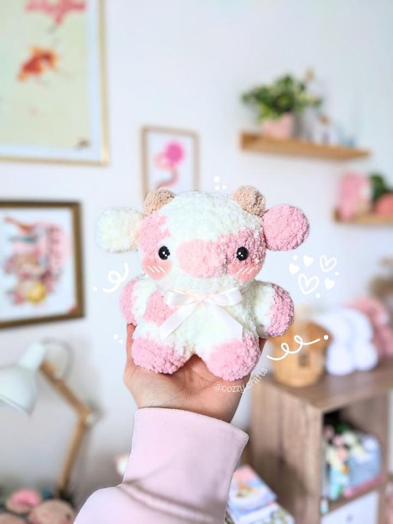 May include: A pink and white crocheted cow plush toy with a white bow. The toy is being held in a hand wearing a pink sweater.