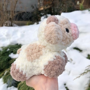 May include: A small, crocheted cow plush toy with brown and white patches. The toy has a pink nose and is held in a hand against a snowy background.