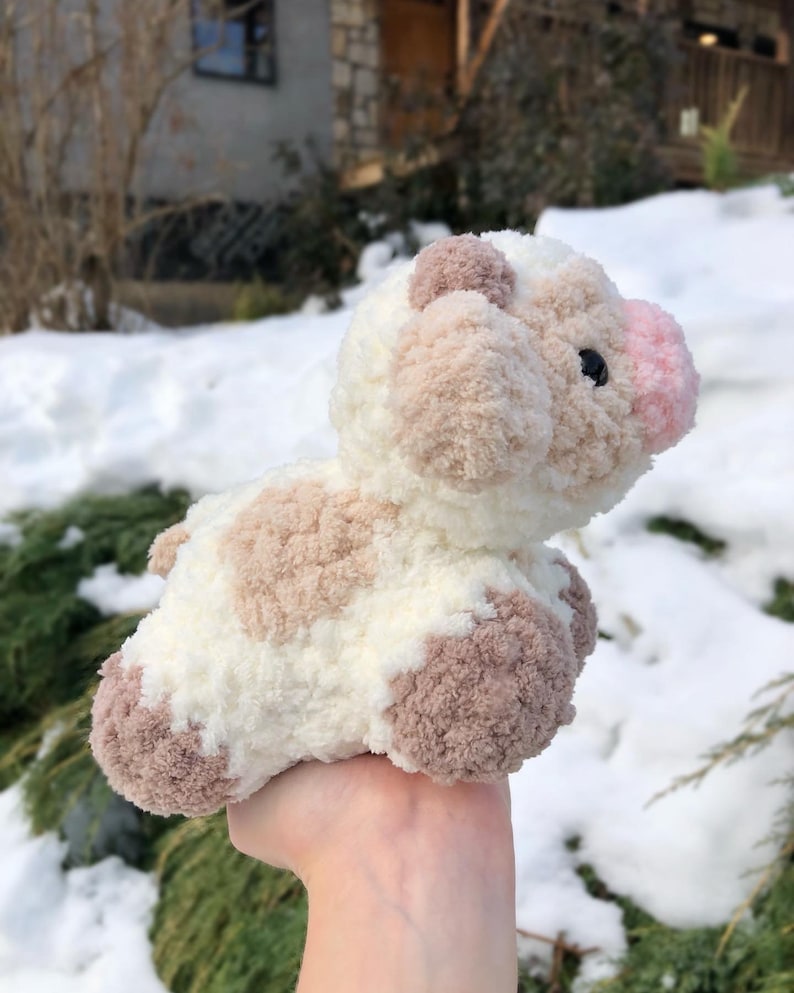May include: A small, crocheted cow plush toy with brown and white patches. The toy has a pink nose and is held in a hand against a snowy background.