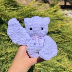 May include: A small, lavender-colored crocheted plush toy shaped like a bear with a pink bow. The bear is being held by a hand in front of a green bush.