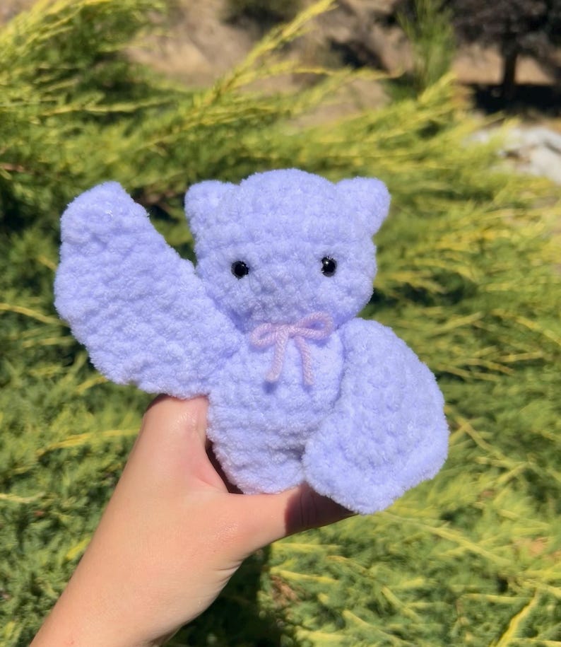 May include: A small, lavender-colored crocheted plush toy shaped like a bear with a pink bow. The bear is being held by a hand in front of a green bush.