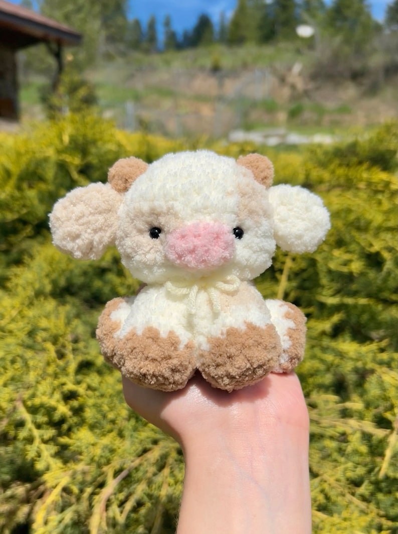 May include: A small crocheted cow plush toy with brown and white fur. The cow has a pink nose and black eyes. The toy is sitting in a person's hand.