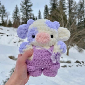 May include: A crocheted plush toy cow with white and purple fur, wearing purple overalls and a pink nose. The cow is holding a purple flower in its hand.
