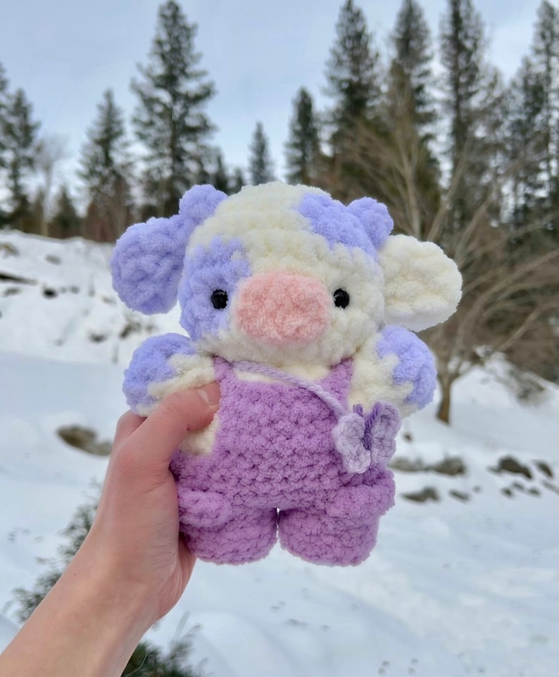 May include: A crocheted plush toy cow with white and purple fur, wearing purple overalls and a pink nose. The cow is holding a purple flower in its hand.