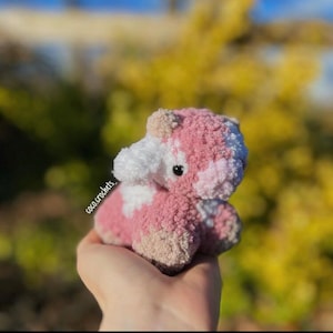 May include: A small pink and white crocheted cow plush toy with black eyes. The toy is being held in a hand.