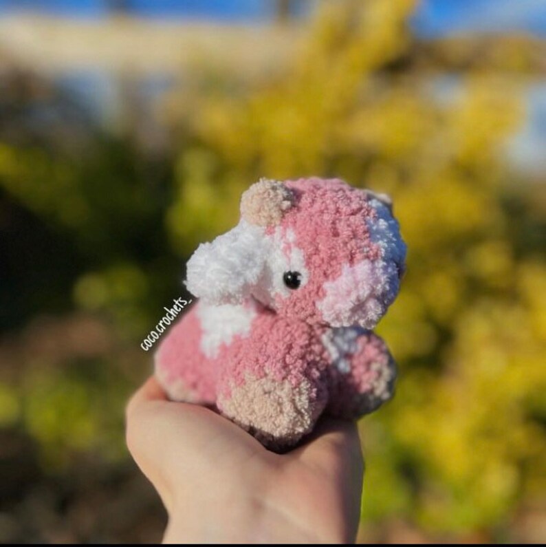 May include: A small pink and white crocheted cow plush toy with black eyes. The toy is being held in a hand.