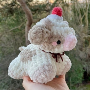 May include: A crocheted plush toy cow with a pink nose, brown eyes, and a red pom-pom hat. The cow is white with brown and pink accents.