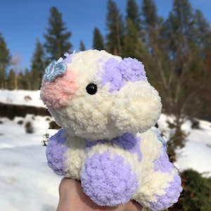 May include: A white and purple crocheted stuffed animal with a pink nose and blue flower accents. The animal is being held in a hand against a snowy background.