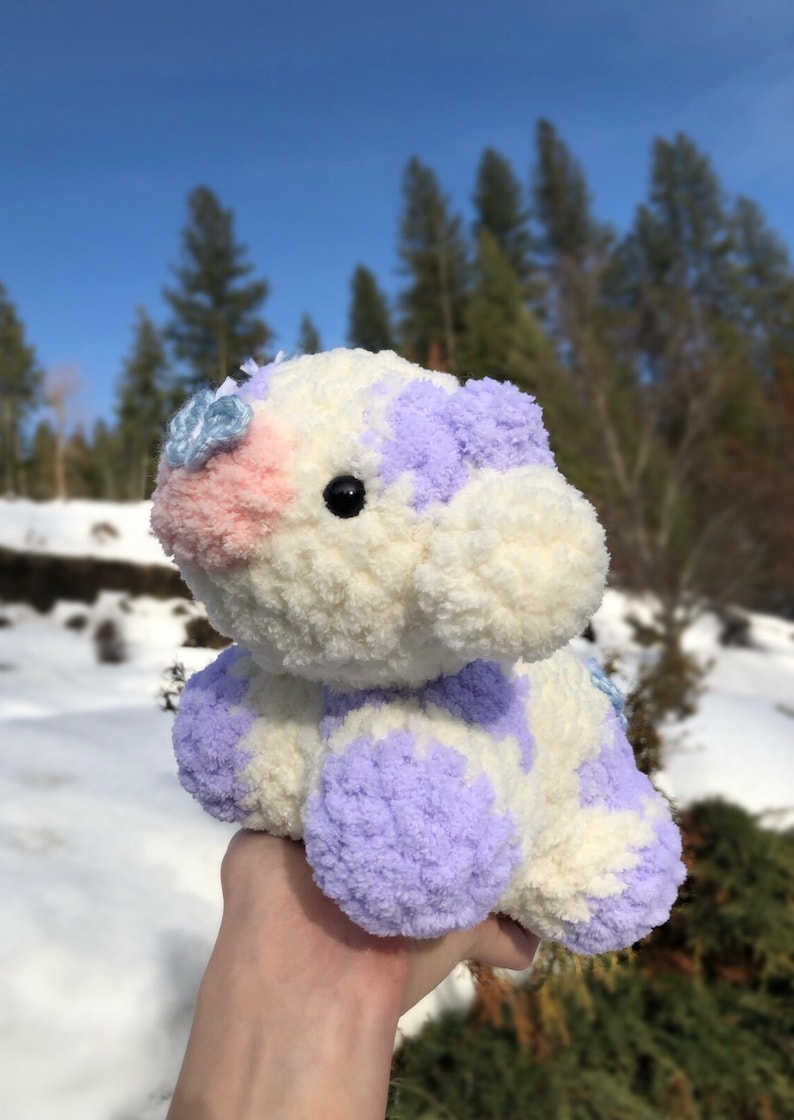 May include: A white and purple crocheted stuffed animal with a pink nose and blue flower accents. The animal is being held in a hand against a snowy background.