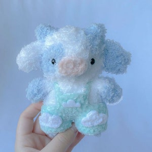 May include: A soft, plush blue and white crocheted cow toy wearing a light green overall with white cloud accents.