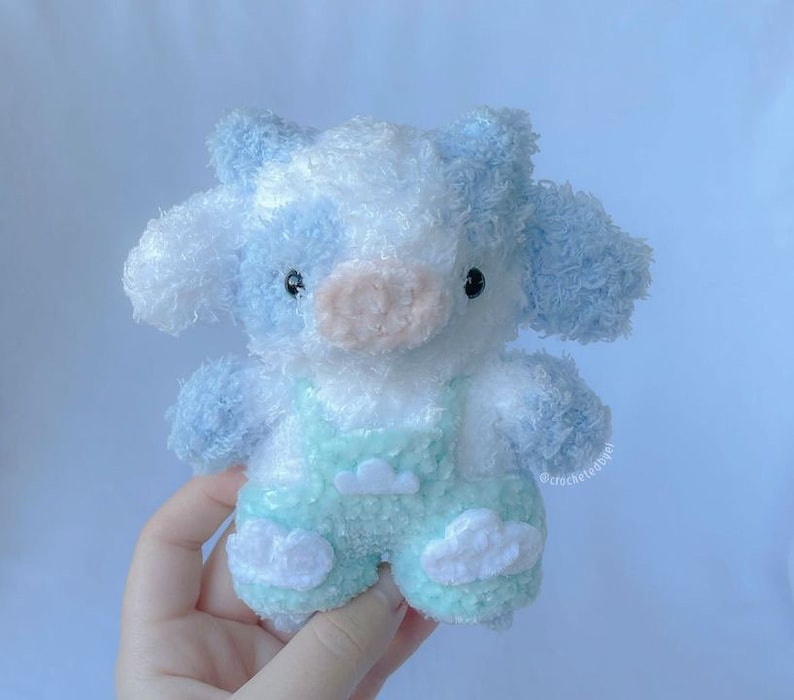May include: A soft, plush blue and white crocheted cow toy wearing a light green overall with white cloud accents.