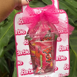 May include: Pink and white cardboard gift bag with a handle, decorated with a repeating pattern of the word "Barbie". The bag contains a clear plastic bag filled with strawberry flavoured candy. The candy is wrapped in individual packets with the brand name "Pop Rocks" printed on them.