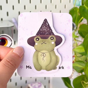 Könnte beinhalten: Ein skurriles Lesezeichen mit einem grünen Frosch, der einen Hexenhut mit Sternen trägt. Der Frosch hat einen hellgrünen Körper, einen dunkelbraunen Hut und einen hellvioletten Hintergrund mit Sternen. Der Text "Magic Mark" ist aufgedruckt.