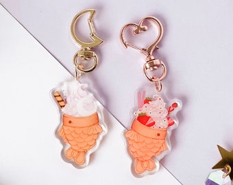 May include: Two acrylic keychains shaped like fish-shaped waffles, one with a wafer and whipped cream, the other with strawberries and a straw. Both have gold-colored clasps. A gold moon and star charm with iridescent crystals is also visible.