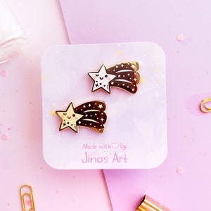 May include: Two enamel pins featuring a shooting star design with a smiling star face. The pins are gold-colored with brown and white accents. The pins are on a light purple card with the text "Made with by Jino's Art".