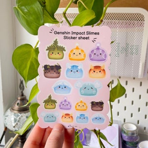 May include: A sticker sheet featuring various Genshin Impact Slimes. The stickers are in a variety of colors, including green, orange, purple, blue, and brown. The sticker sheet has a pink scalloped edge and the text "Genshin Impact Slimes Sticker sheet".