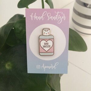 K&ouml;nnte beinhalten: Eine Anstecknadel mit einem Handdesinfektionsmittel-Flaschendesign mit einem l&auml;chelnden Gesicht und einem Herz mit der Aufschrift "Hand Sanitizer". Die Anstecknadel ist rosa, wei&szlig; und goldfarben, mit dem Text "Hand Sanitizer" im Herzen. Der Hintergrund ist ein Farbverlauf aus Rosa und Blau.