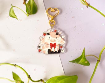 Könnte beinhalten: Ein klarer Acryl-Schlüsselanhänger mit einer Cartoon-Kuh mit einem rosa-weißen Fleckenmuster. Die Kuh hat eine rote Schleife, kleine schwarze Hörner und ist von Erdbeeren und Blumen umgeben. Der Schlüsselanhänger hat einen goldenen herzförmigen Verschluss.