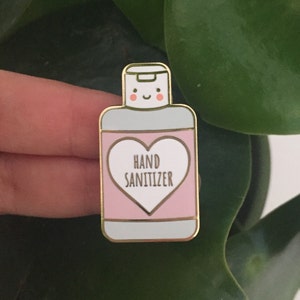K&ouml;nnte beinhalten: Ein Emaille-Anstecknadel in Form einer Handdesinfektionsmittelflasche. Die Nadel ist hellblau, rosa und goldfarben mit den Worten "HAND SANITIZER" in Herzform. Die Flasche hat ein l&auml;chelndes Gesicht.