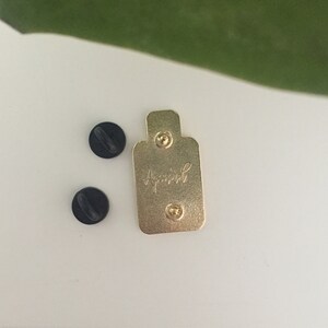 K&ouml;nnte beinhalten: Ein goldfarbener Emaille-Pin mit dem Wort "April" in Schreibschrift. Zwei schwarze Pin-R&uuml;ckseiten befinden sich neben dem Pin. Der Pin ist rechteckig mit abgerundeten Kanten und zwei Pin-Pfosten.