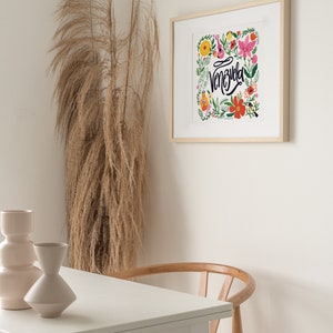 May include: A framed art print featuring the word "Venezuela" in cursive, surrounded by colorful flowers and foliage. The print is in a light wood frame, hanging on a white wall. A table with two vases and a wooden chair are in the foreground.