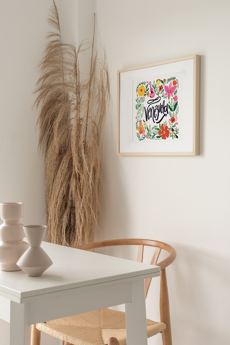 May include: A framed art print featuring the word "Venezuela" in cursive, surrounded by colorful flowers and foliage. The print is in a light wood frame, hanging on a white wall. A table with two vases and a wooden chair are in the foreground.