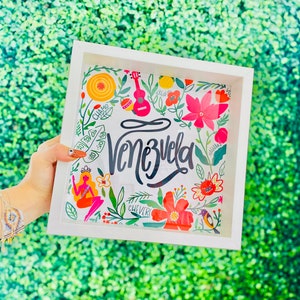 May include: A white framed art print featuring the word "Venezuela" in elegant script, surrounded by colorful floral and cultural illustrations. The artwork includes a ukulele, flowers, and a mermaid, all set against a green background.