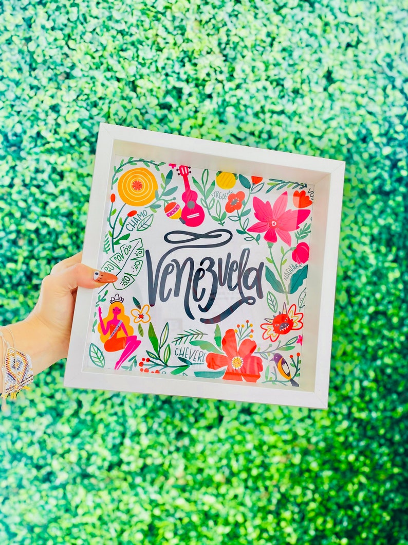 May include: A white framed art print featuring the word "Venezuela" in elegant script, surrounded by colorful floral and cultural illustrations. The artwork includes a ukulele, flowers, and a mermaid, all set against a green background.