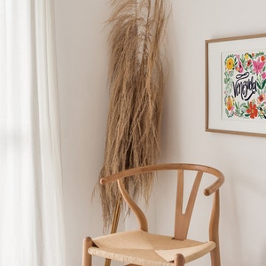 May include: A framed art print with the word "Venezuela" in cursive, surrounded by colorful flowers. A wooden chair with a woven seat sits in front of a white wall. Tall, dried ornamental grass adds a natural element to the scene.