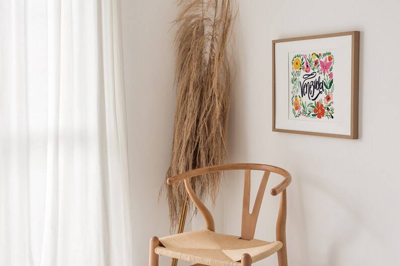 May include: A framed art print with the word "Venezuela" in cursive, surrounded by colorful flowers. A wooden chair with a woven seat sits in front of a white wall. Tall, dried ornamental grass adds a natural element to the scene.