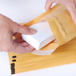 Peut inclure: Une personne emballe une boîte blanche dans une enveloppe matelassée brune. L'enveloppe fait partie d'une pile d'enveloppes.