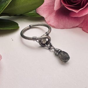 Puede incluir: Un anillo de plata hecho a mano con un diseño único. El anillo presenta una banda circular con un detalle en forma de nudo y una piedra colgante en forma de lágrima. La piedra está envuelta en alambre de plata, agregando textura. Fondo blanco.