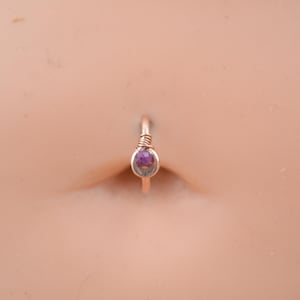 May include: A close-up of a belly button ring with a small purple gemstone. The ring is made of gold-toned wire.