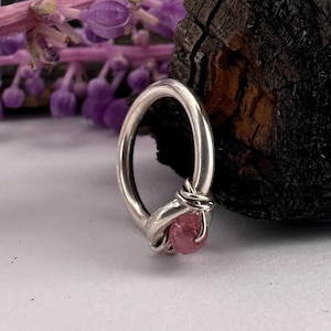 May include: A silver ring with a pink gemstone. The ring features a smooth, polished band that curves into a loop. The gemstone is a round, pink stone, securely held in place by a wire wrap. The ring is set against a soft, white background.
