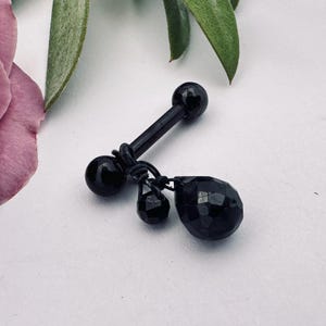 May include: Black barbell body jewelry with a teardrop charm and two smaller charms. The jewelry is made of black metal and has a polished finish. The teardrop charm has a faceted design.
