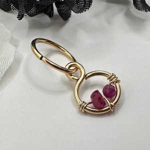 May include: A gold-colored earring with a figure-eight design. Two small, rough-cut, ruby-colored stones are held within the lower loop of the earring. The earring is on a white surface with a black floral accent in the background.