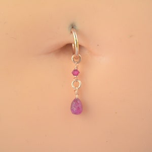 Peut inclure: Un piercing de nombril avec un charme suspendu orné d'une pierre précieuse rose en forme de larme et de deux autres pierres précieuses roses plus petites.