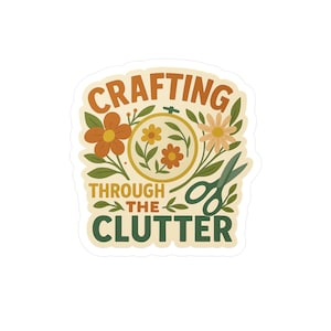 May include: A sticker with the text "CRAFTING THROUGH THE CLUTTER" in orange and green. The design includes flowers, leaves, a needlepoint hoop, and scissors. The sticker has a cream-colored background.