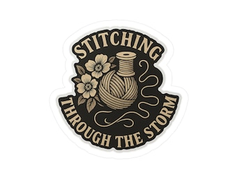 Könnte beinhalten: Ein Aufkleber mit dem Text "Stitching Through The Storm" in einem geschwungenen Banner. Das Design zeigt eine Garnspule, einen Wollknäuel und florale Elemente in Beige und Schwarz.