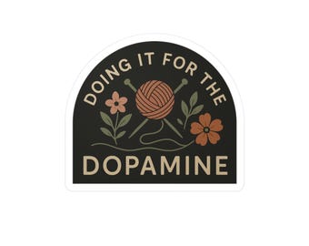 Könnte beinhalten: Aufkleber mit schwarzem Hintergrund und weißem Rand. Der Text "DOING IT FOR THE DOPAMINE" ist halbkreisförmig angeordnet. Er enthält auch einen Wollknäuel, Stricknadeln und florale Elemente.