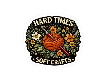 Könnte beinhalten: Ein Aufkleber mit dem Text "HARD TIMES SOFT CRAFTS" in Weiß, der über und unter einem zentralen Bild angeordnet ist. Das Bild zeigt einen orangefarbenen Wollknäuel mit einem roten Häkelhaken, umgeben von Blumen und Blättern in Grün, Gelb und Orange.