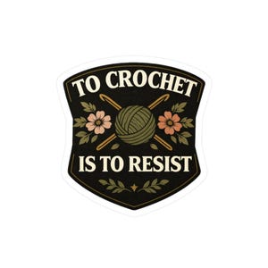 May include: A black sticker with the text "TO CROCHET IS TO RESIST" in white. The design includes a ball of yarn, two crochet hooks, and floral accents in pink and green. The sticker has a shield shape.
