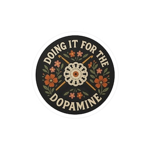 以下が含まれることがあります： クリーム色の文字で「DOING IT FOR THE DOPAMINE」と書かれた丸いステッカー。黒い背景にオレンジとクリーム色の花、2本の編み棒、中央にクリーム色の編み花の模様があります。