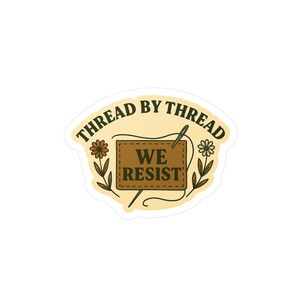 May include: A sticker featuring a tan background with a brown rectangle at the centre. The rectangle displays the words "WE RESIST" in white. The words "THREAD BY THREAD" are arched above. Floral designs are on either side.