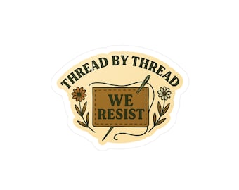 Könnte beinhalten: Ein Aufkleber mit beigefarbenem Hintergrund und einem braunen Rechteck in der Mitte. Das Rechteck zeigt die weißen Worte "WE RESIST". Die Worte "THREAD BY THREAD" sind bogenförmig darüber angeordnet. Blumendesigns befinden sich auf beiden Seiten.