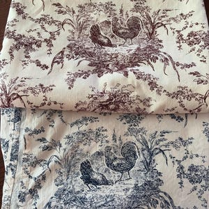 May include: Two folded pieces of fabric featuring a toile pattern. The top piece has a dark red print on a cream background, while the bottom piece has a dark blue print on a cream background. Both display illustrations of roosters and hens.